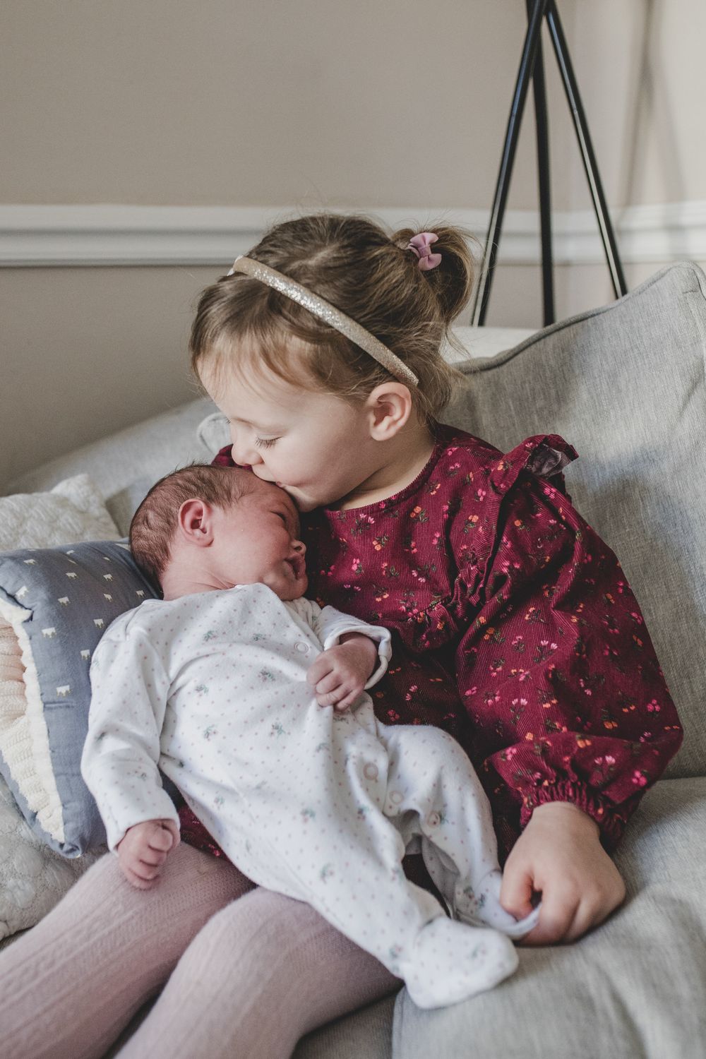 A young child in a burgundy dress tenderly kisses a baby wearing white while sitting together on a grey couch.