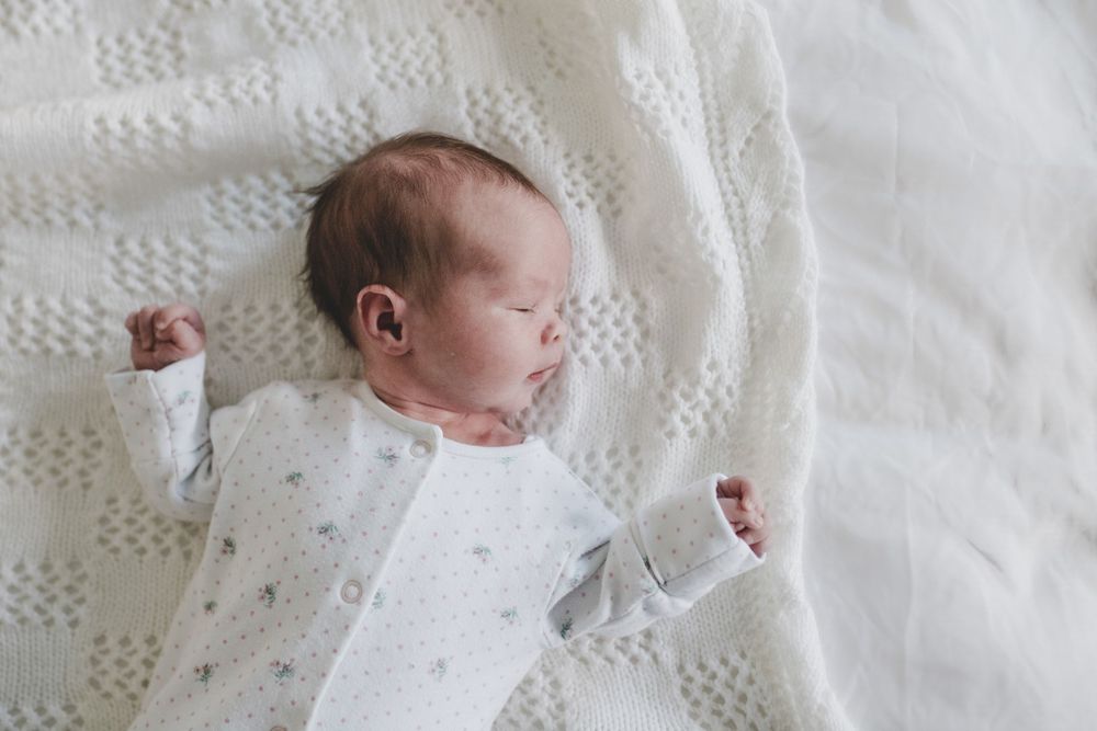 Sleeping newborn wearing white sleepwear rests peacefully on textured white blanket.