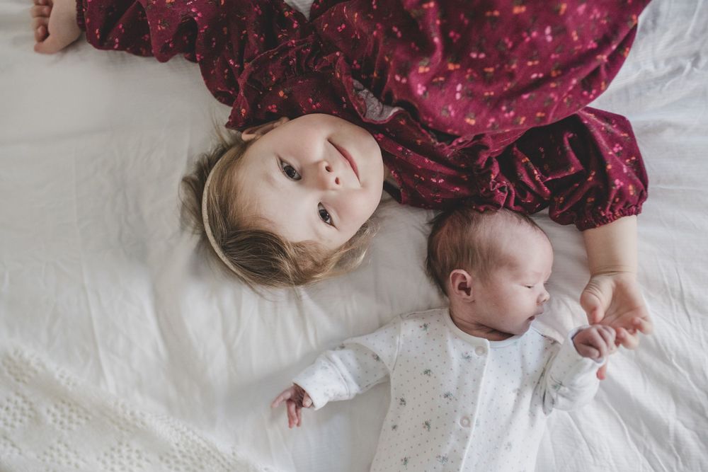 A parent in a red dress lies next to a sleeping baby in white on a white bed.