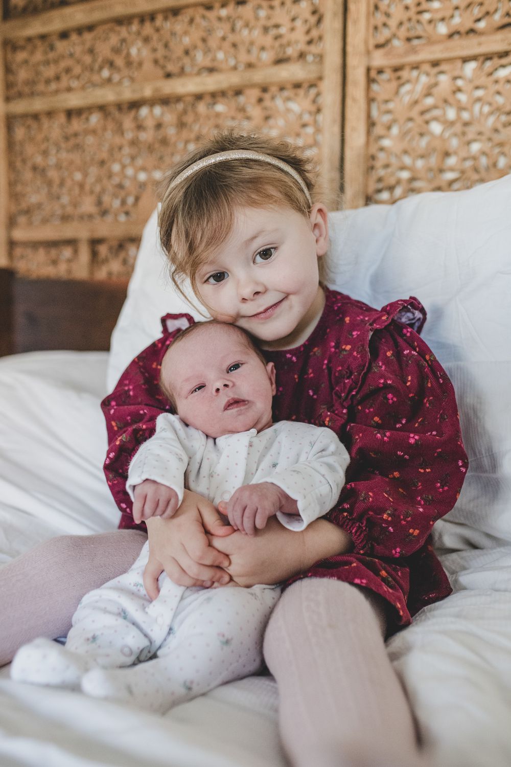 Two children sit together on white bedding, with the older one wearing a burgundy patterned dress holding a baby in white.