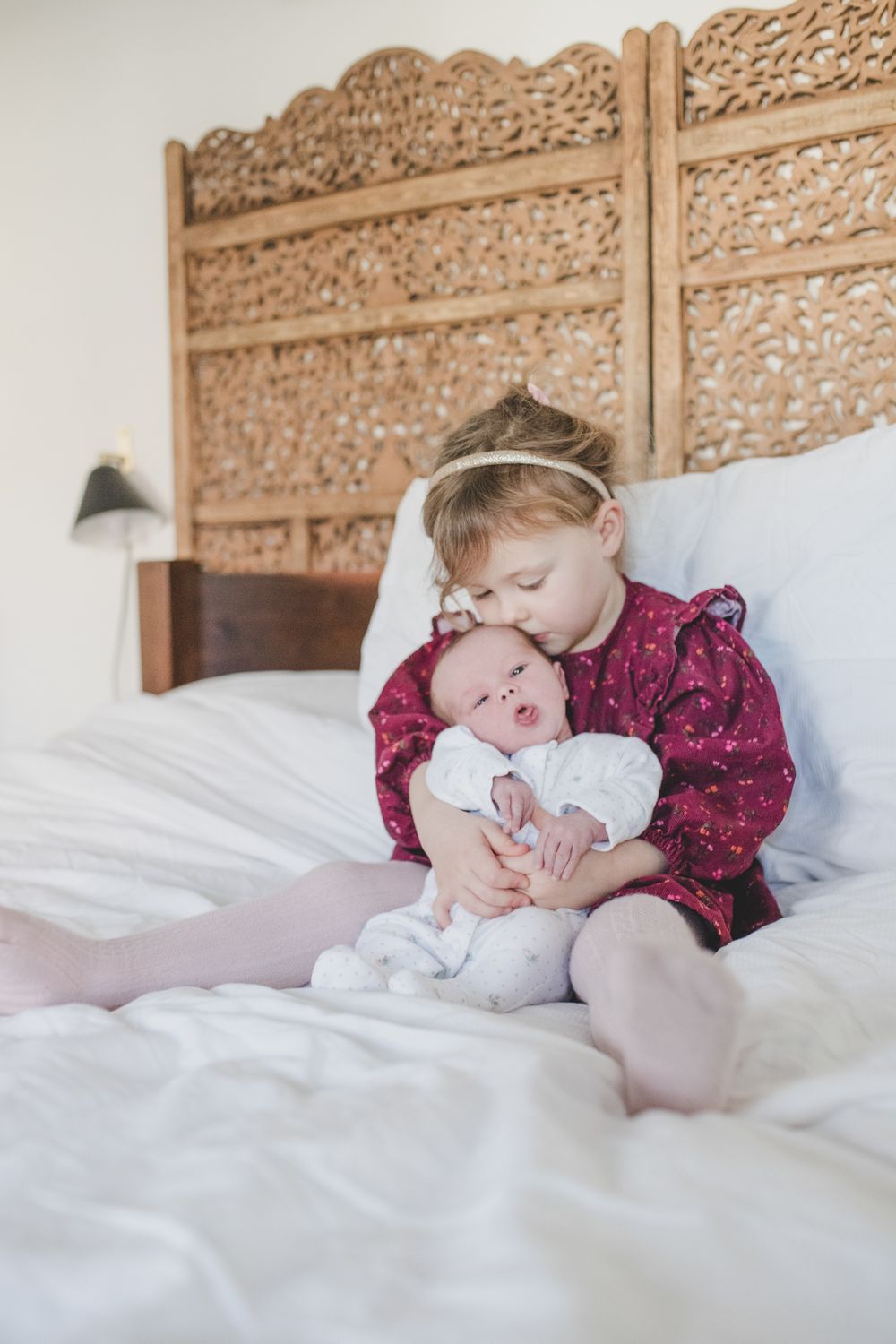 A sweet moment of comfort and bonding on a white bed with an ornate wooden headboard in the background.