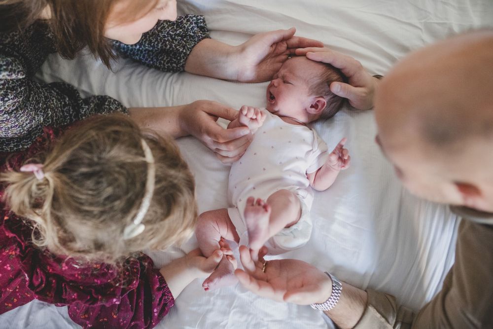 Family gathered around newborn baby lying on white bed wearing pink outfit with others reaching to touch and interact.