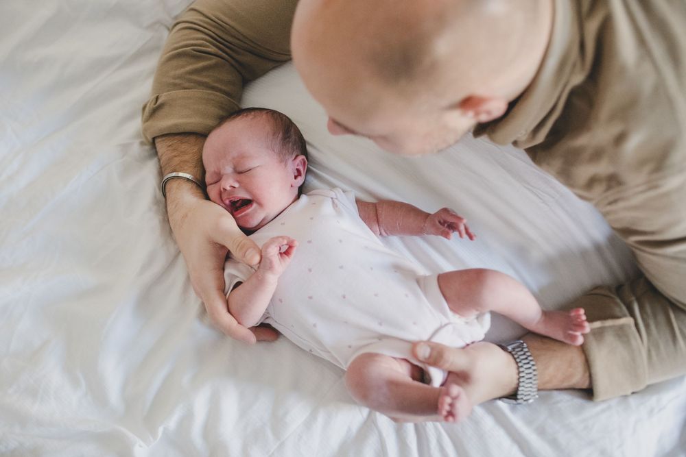 Tender moment showing a newborn baby in white onesie being cradled on soft bedding.