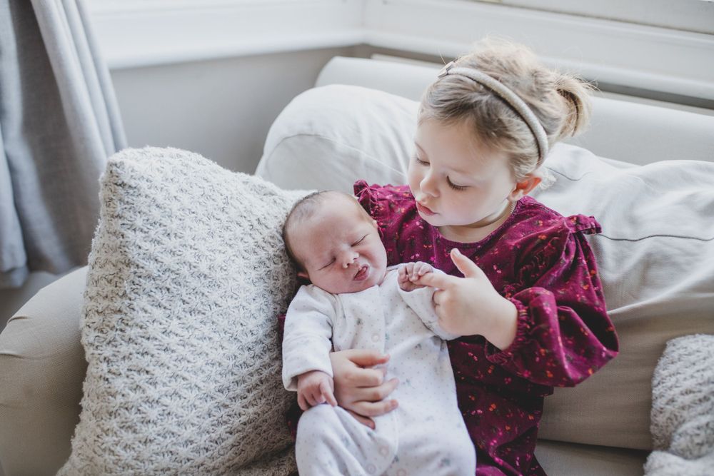 A young child wearing a burgundy top sits on a couch while gently holding a crying baby near white pillows.