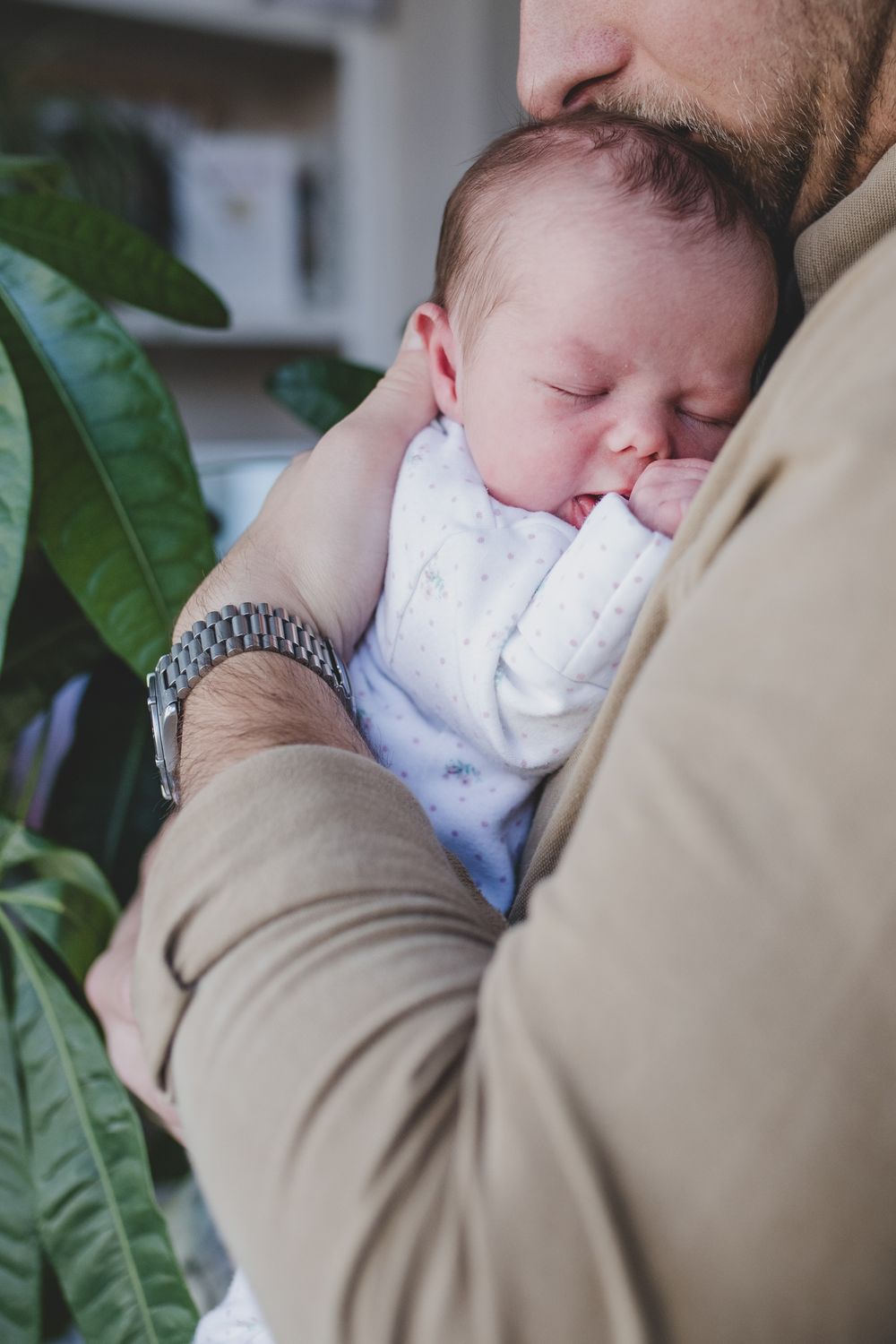 A sleeping newborn wearing white clothing rests peacefully against someone in a tan jacket near green foliage.