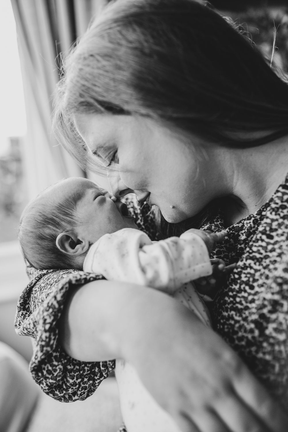 A tender black and white photo of a mother cradling and kissing her newborn baby in a floral dress.