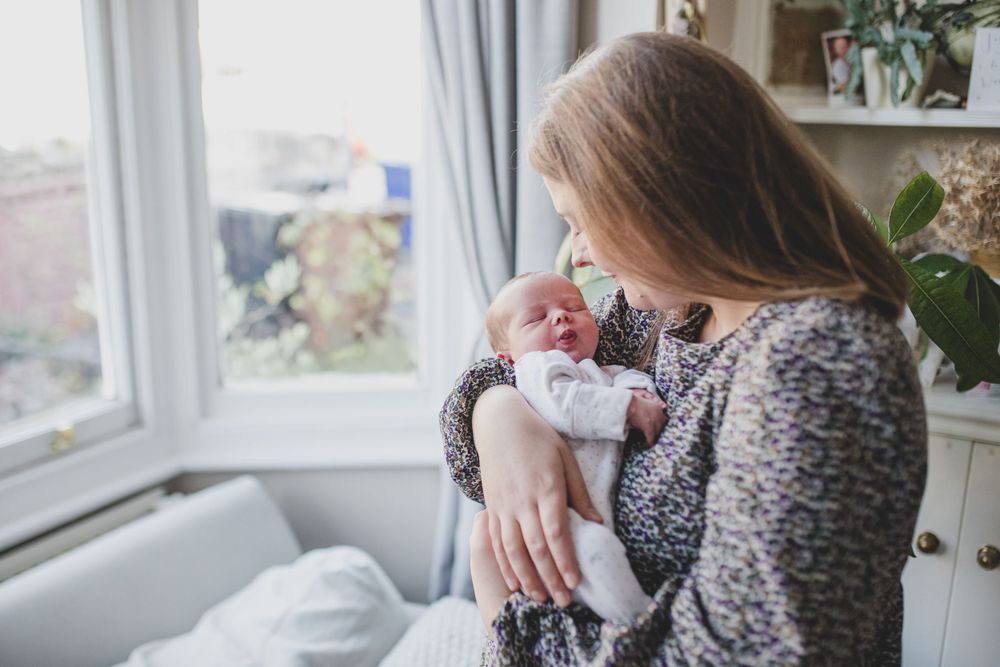 A parent in a gray sweater holds and gazes lovingly at their sleeping newborn baby near a bright window.