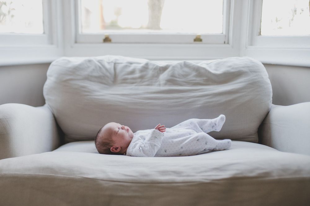 Sleeping infant in white onesie rests on plush light-colored couch near a large window.
