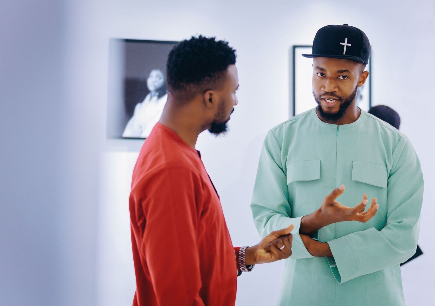 Two men stand in a room having a conversation, one wearing a red shirt and another in mint green scrubs.