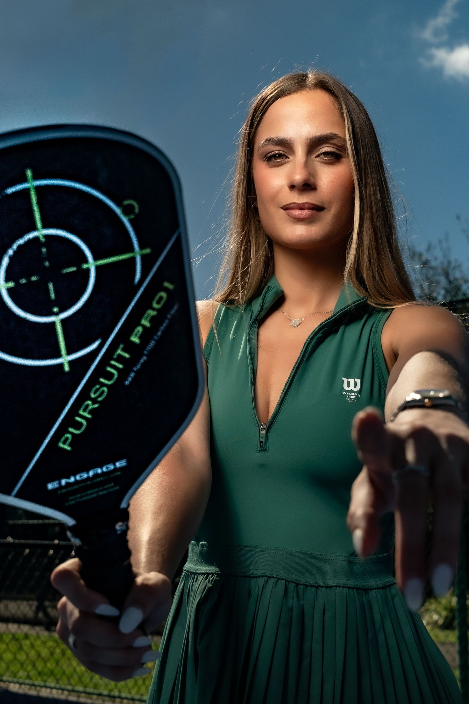 Athletic model in green tennis dress showcases modern tennis tracking device against blue sky.