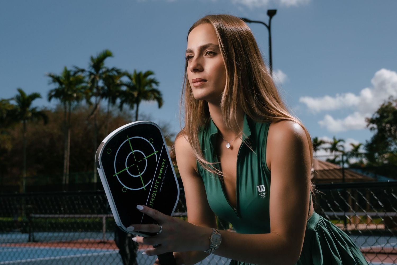 Tennis player holding electronic tracking device on outdoor court with palm trees in background.