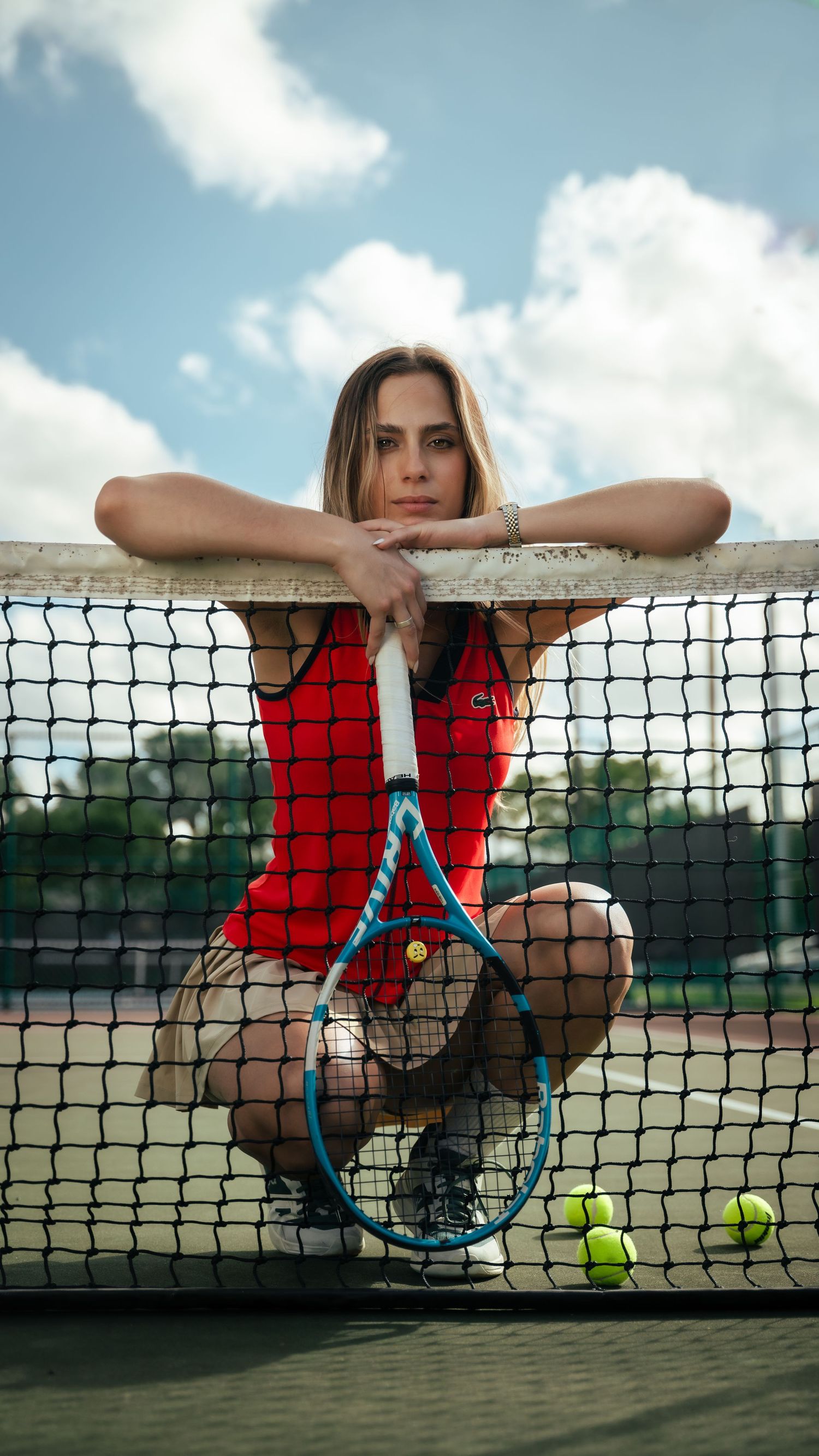 Tennis player in red outfit leans on court net with racquet and tennis balls.