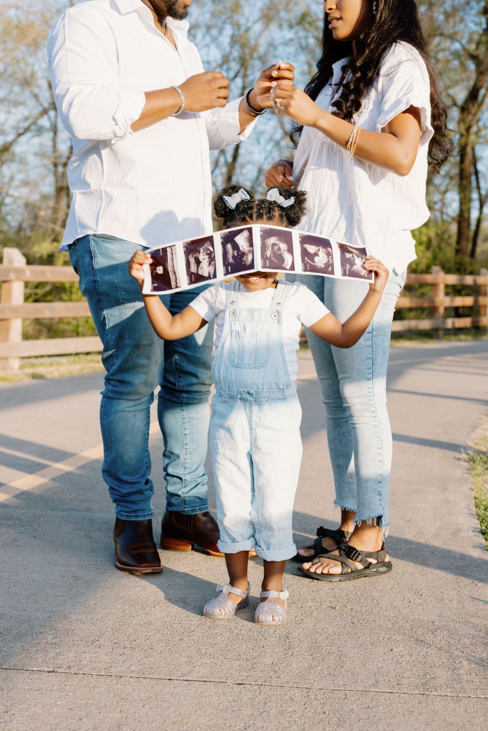 Family stands outdoors holding ultrasound pictures with child between them wearing white overalls.