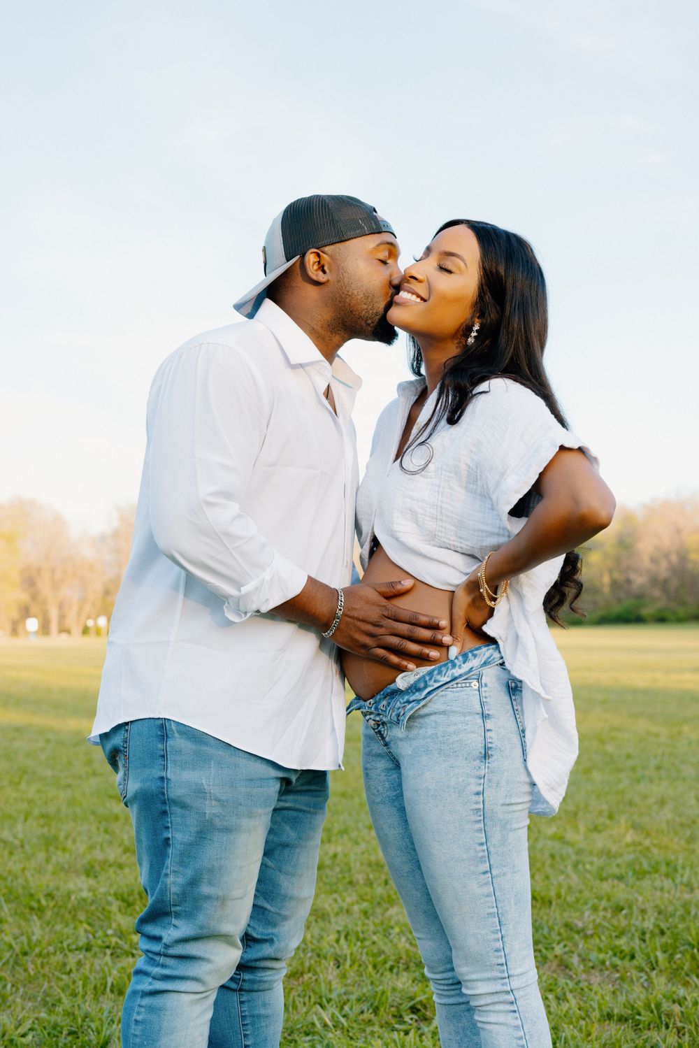 A couple shares a tender kiss while standing in a green field during golden hour wearing casual denim and white tops.