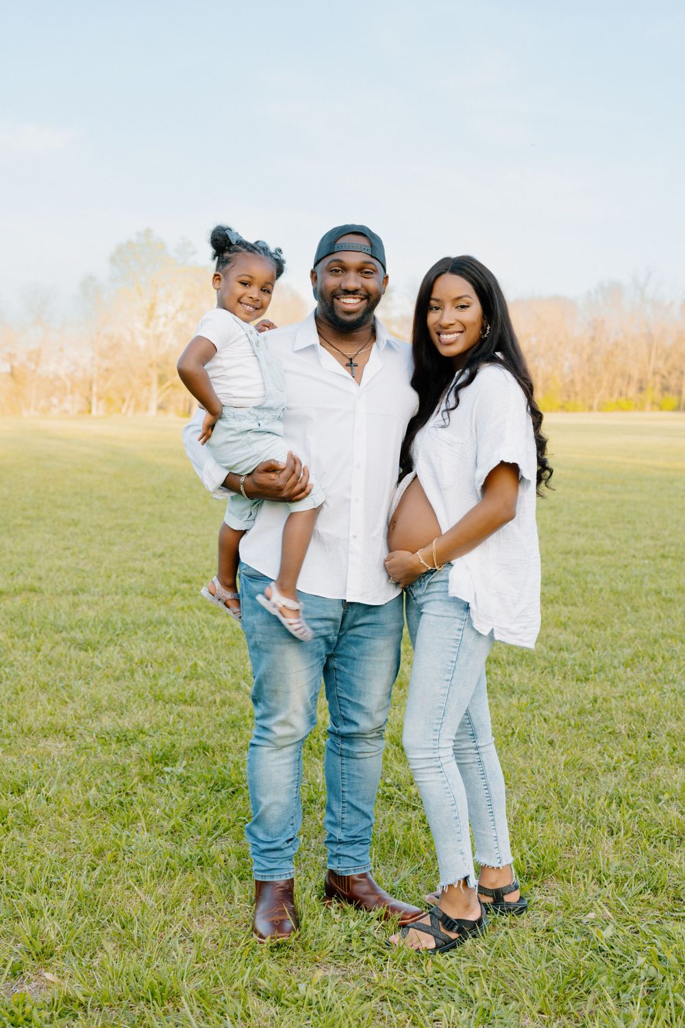 A family wearing matching white tops and jeans poses together outdoors in a grassy field with golden fall foliage behind them.