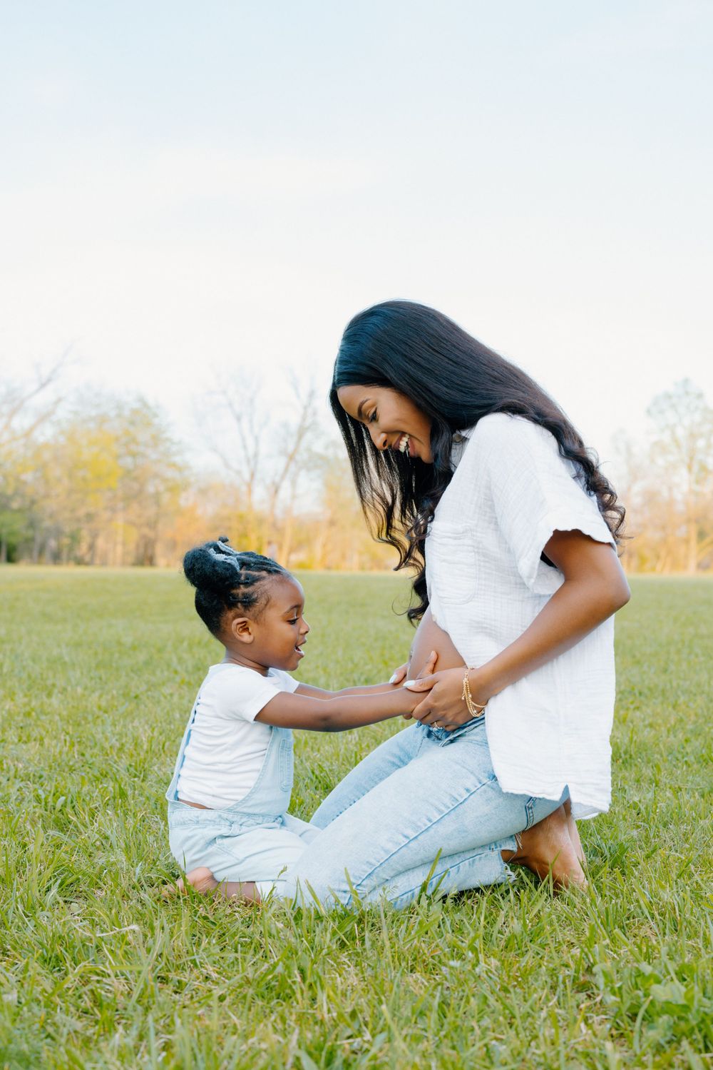 Mother and child wearing coordinated white shirts share a tender moment together while sitting in a grassy field.