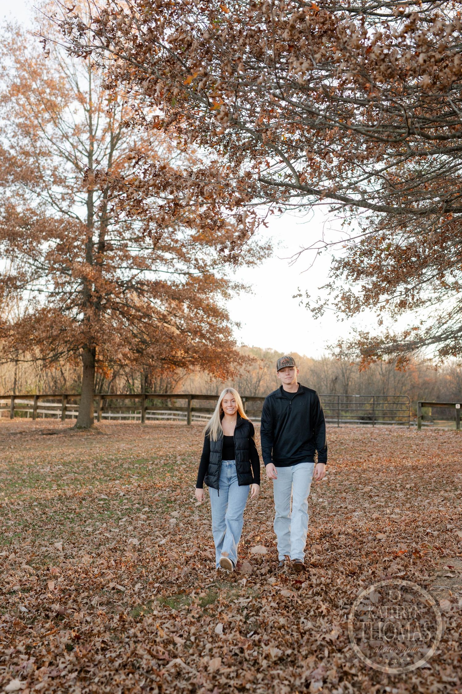 older siblings walks together on fallen leaves with autumn trees and wooden fencing in background.