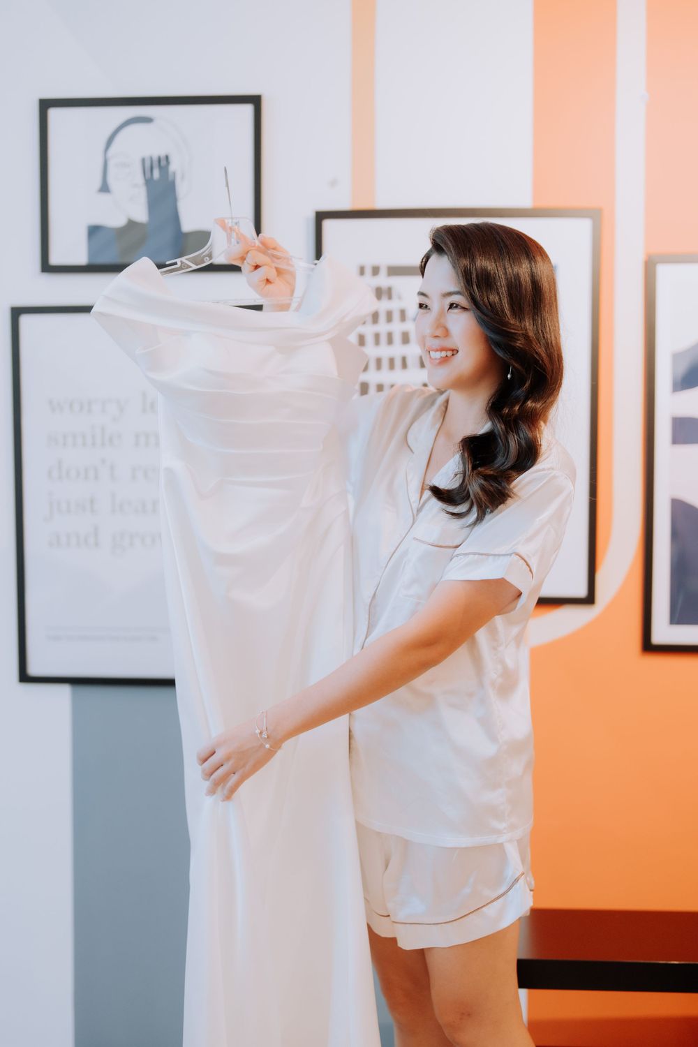 A person in white pajamas holds up a white dress while smiling in a room with framed artwork on orange walls.