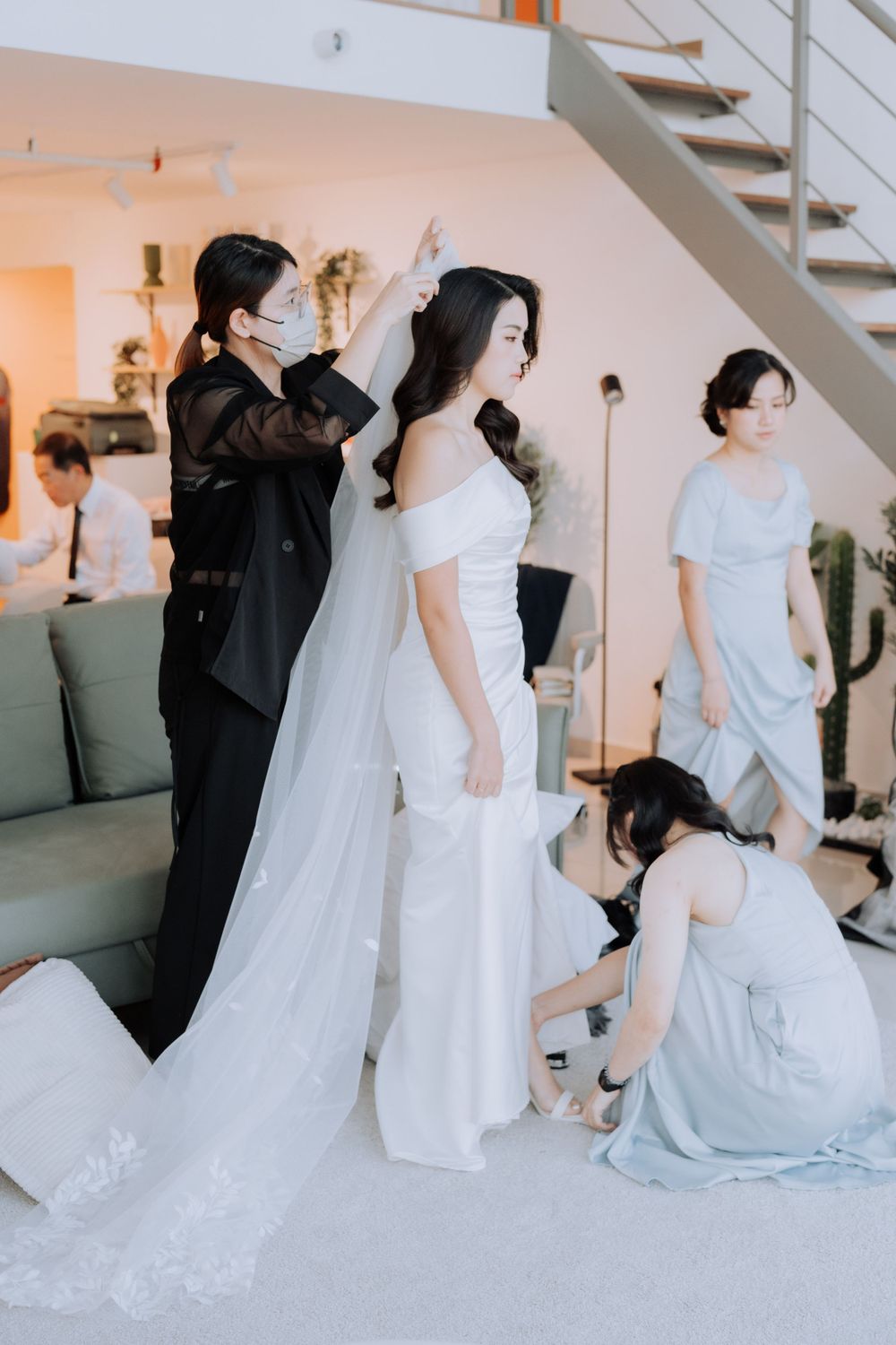 Wedding preparations scene showing a bride in a flowing white dress getting final adjustments in a modern interior setting.