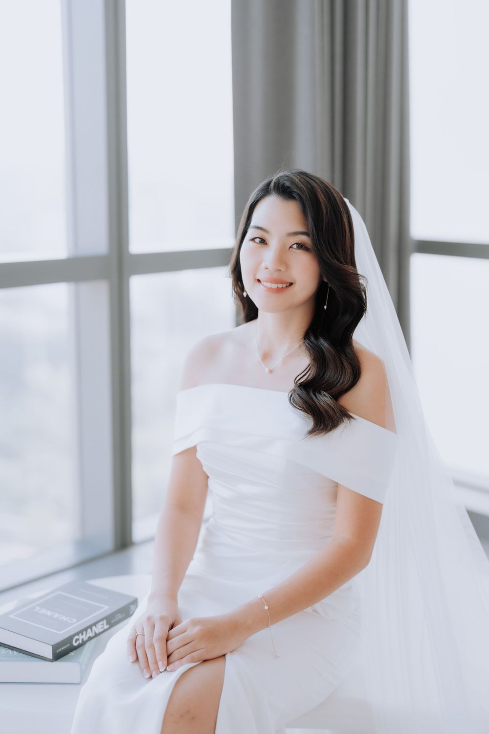 Elegant bride in white off-shoulder wedding dress sits gracefully by a bright window.