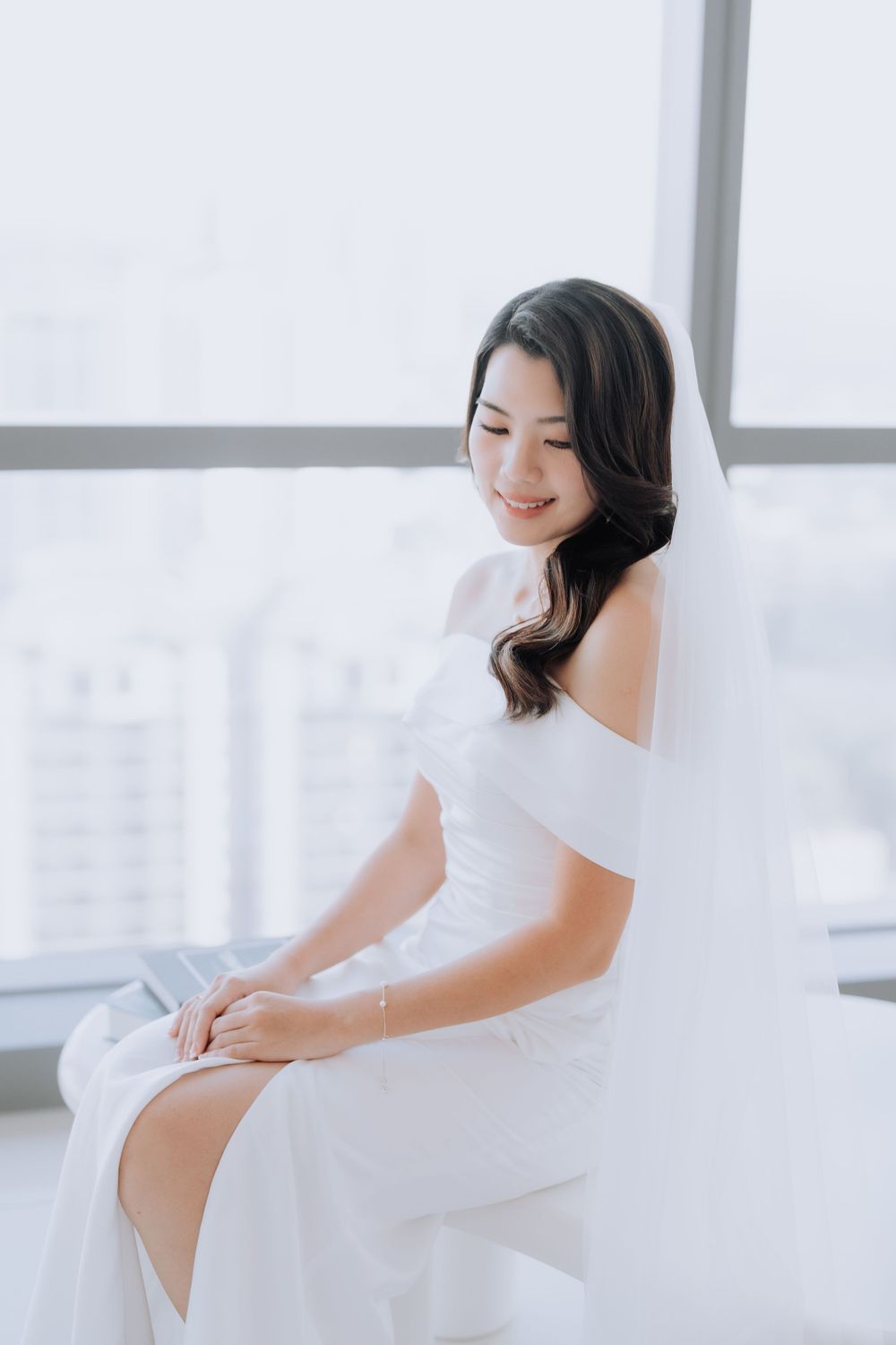 Woman in white off-shoulder dress sits by window smiling with soft, ethereal lighting.