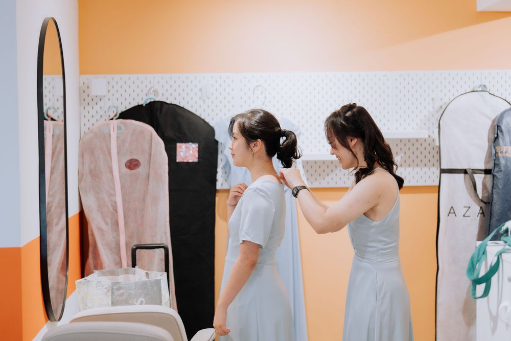 Two people in gray dresses help each other get ready in a dressing room with orange walls.