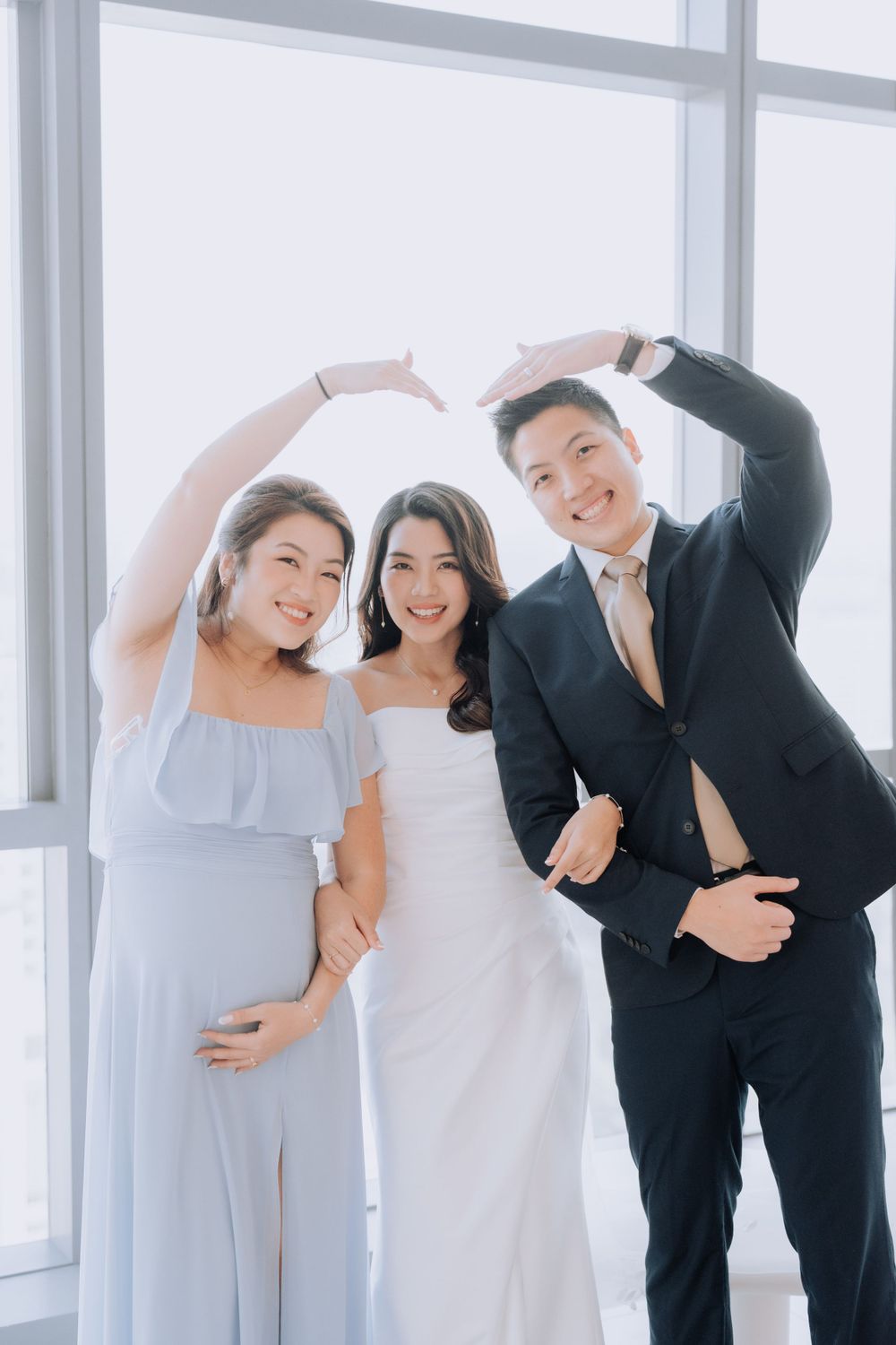 Joyful wedding party members pose together making heart shapes with their hands against bright window.