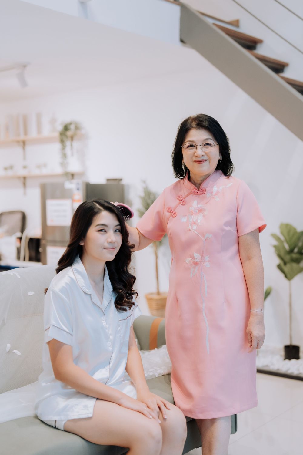 Two women in traditional Chinese-style dresses, one in pink and one in white, pose together in a bright interior setting.