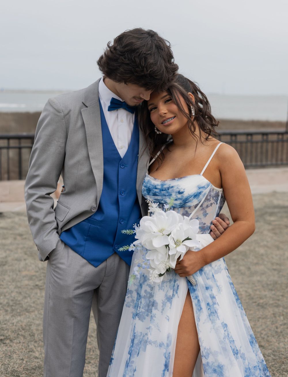 A couple in formal attire embrace at a beach wedding, with her wearing a blue and white floral dress and him in gray suit.