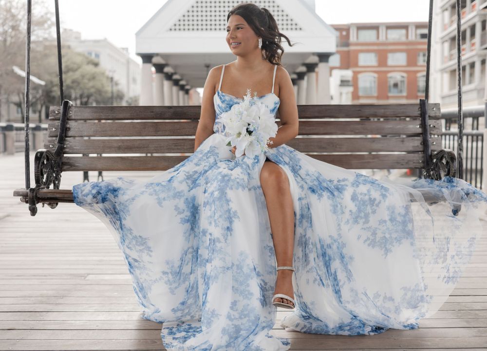 A flowing sky blue and white floral gown with spaghetti straps billows in the breeze on a wooden boardwalk.