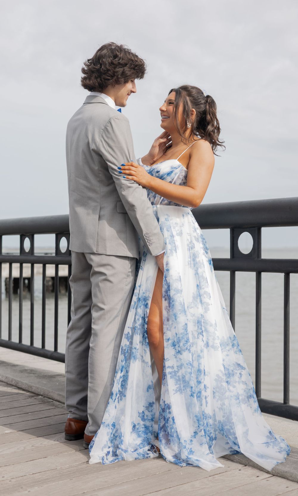 A couple in formal attire share an intimate moment on a boardwalk, with her wearing a blue and white floral gown.