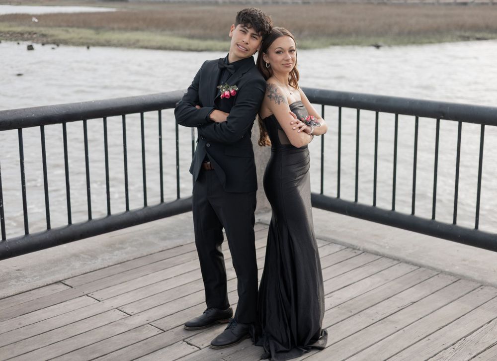 A couple dressed in formal attire poses back-to-back on a wooden deck overlooking water.
