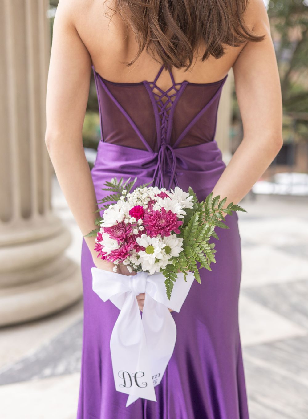 A person in a purple formal dress holds a bouquet of white and pink flowers with ferns against classical columns.