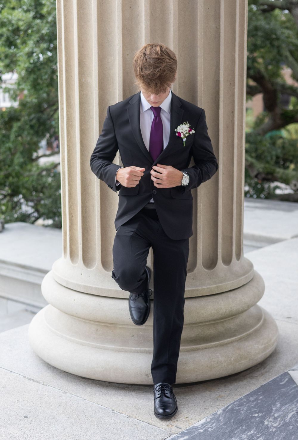 A well-dressed person in a black suit with purple tie adjusts their jacket while standing by a classical column.