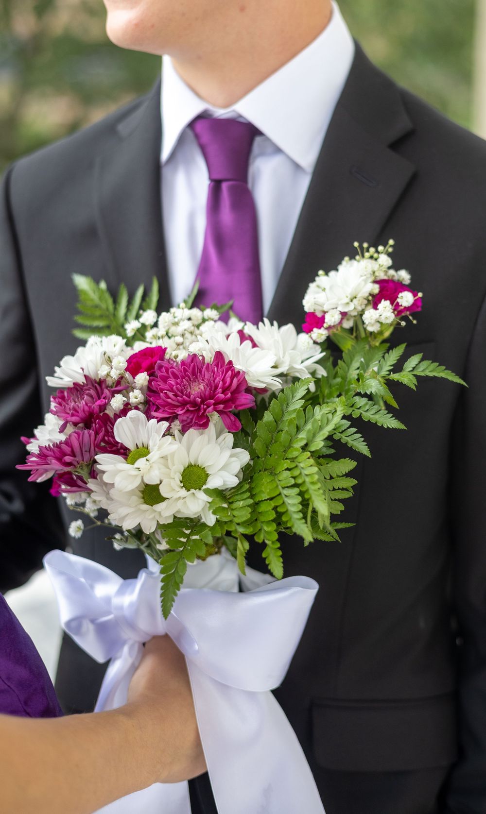 A formal boutonniere featuring purple and white flowers with ferns against a black suit and purple tie.