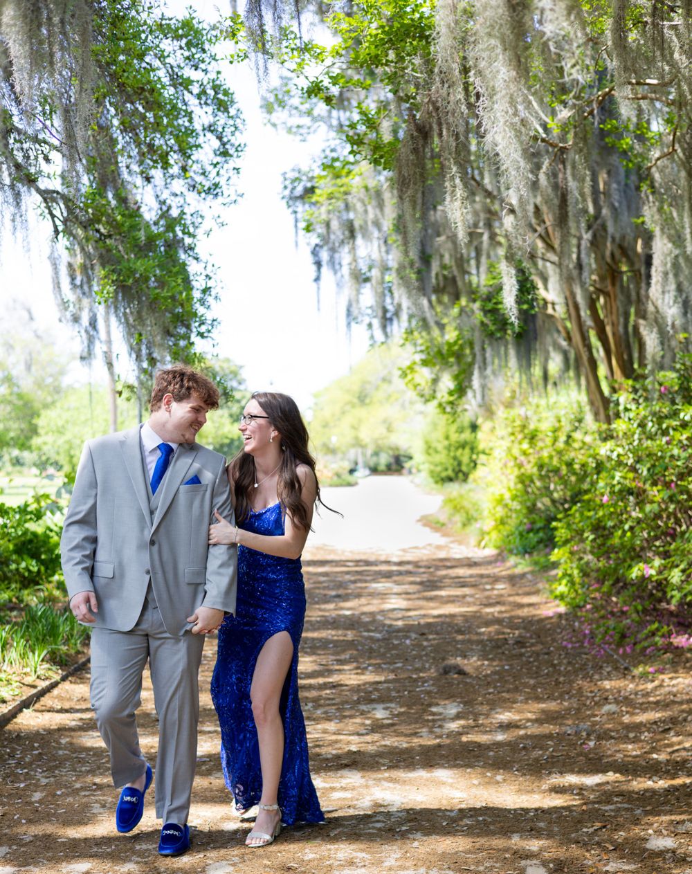 A couple in formal attire walks together on a tree-lined path with Spanish moss hanging overhead in a romantic setting.