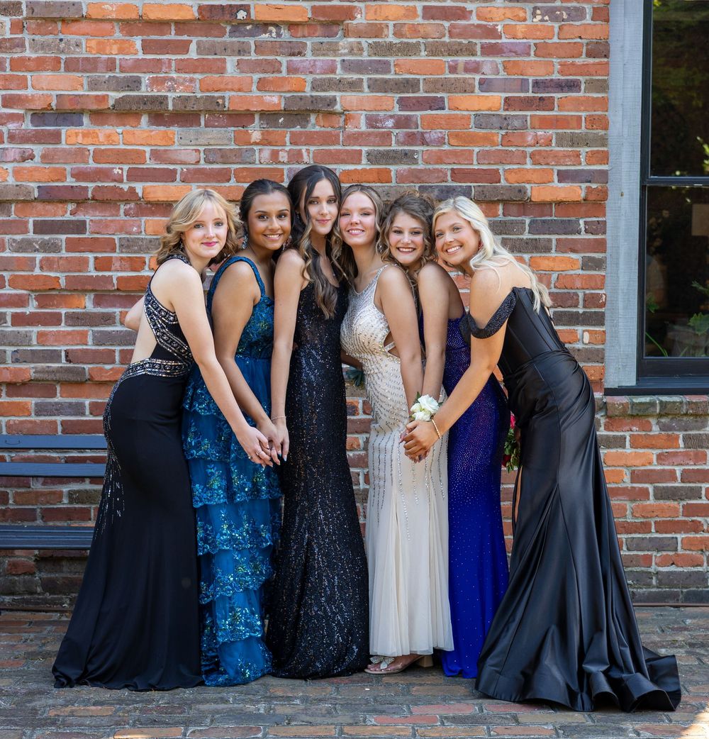 A group of friends in formal evening gowns pose together against a brick wall during a special occasion.