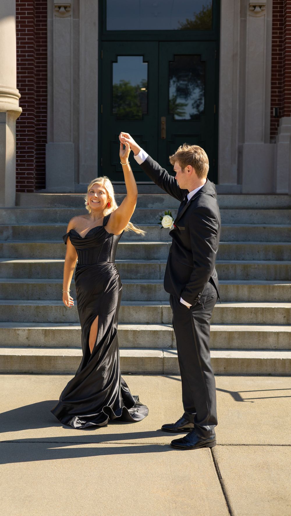 Couple in formal black attire dancing on steps of brick building in bright sunlight, with elegant twirling pose.