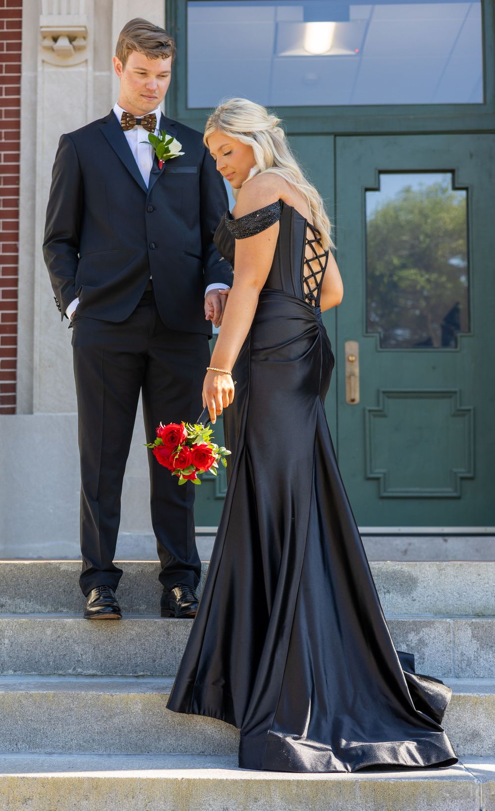 A couple in formal attire poses on stone steps, with her wearing a black gown and holding red flowers.