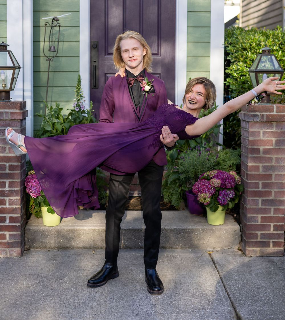 A couple shares a joyful moment on a front porch, with the person wearing a flowing purple dress being playfully lifted.