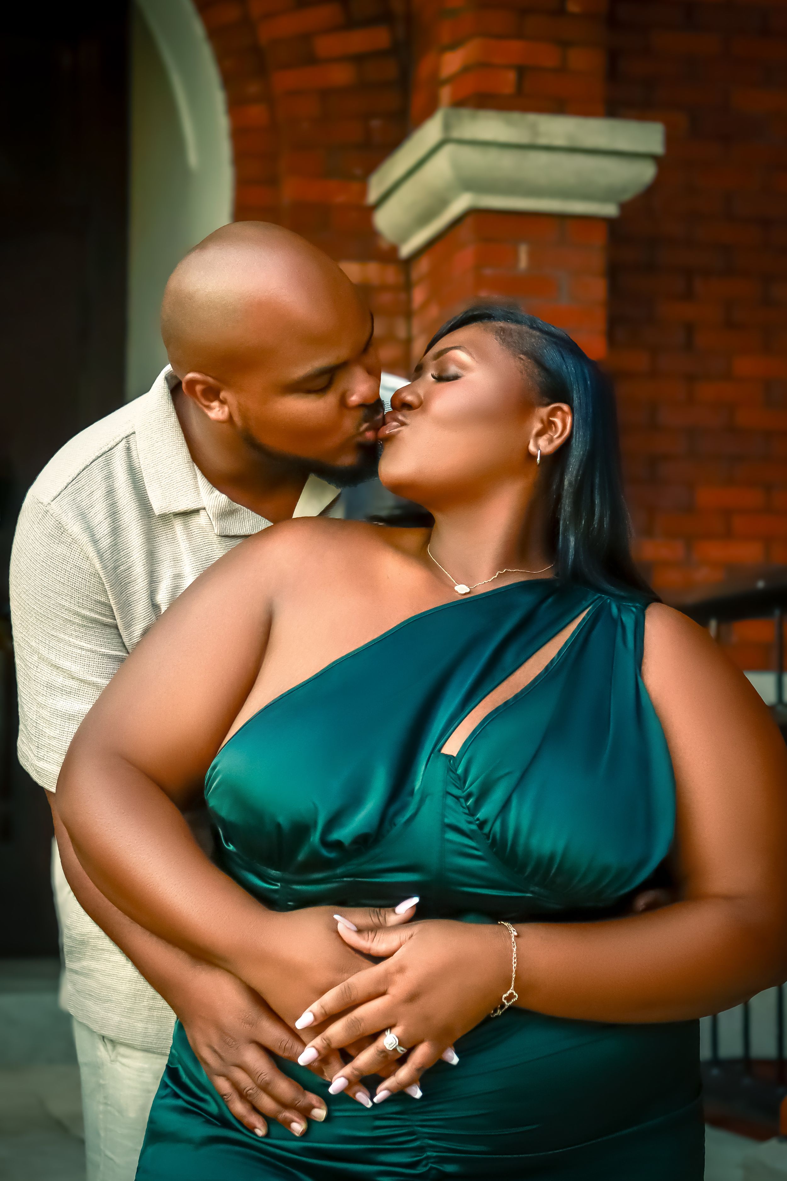 A romantic moment captured between a couple embracing in front of a brick wall, with one wearing an elegant teal dress.