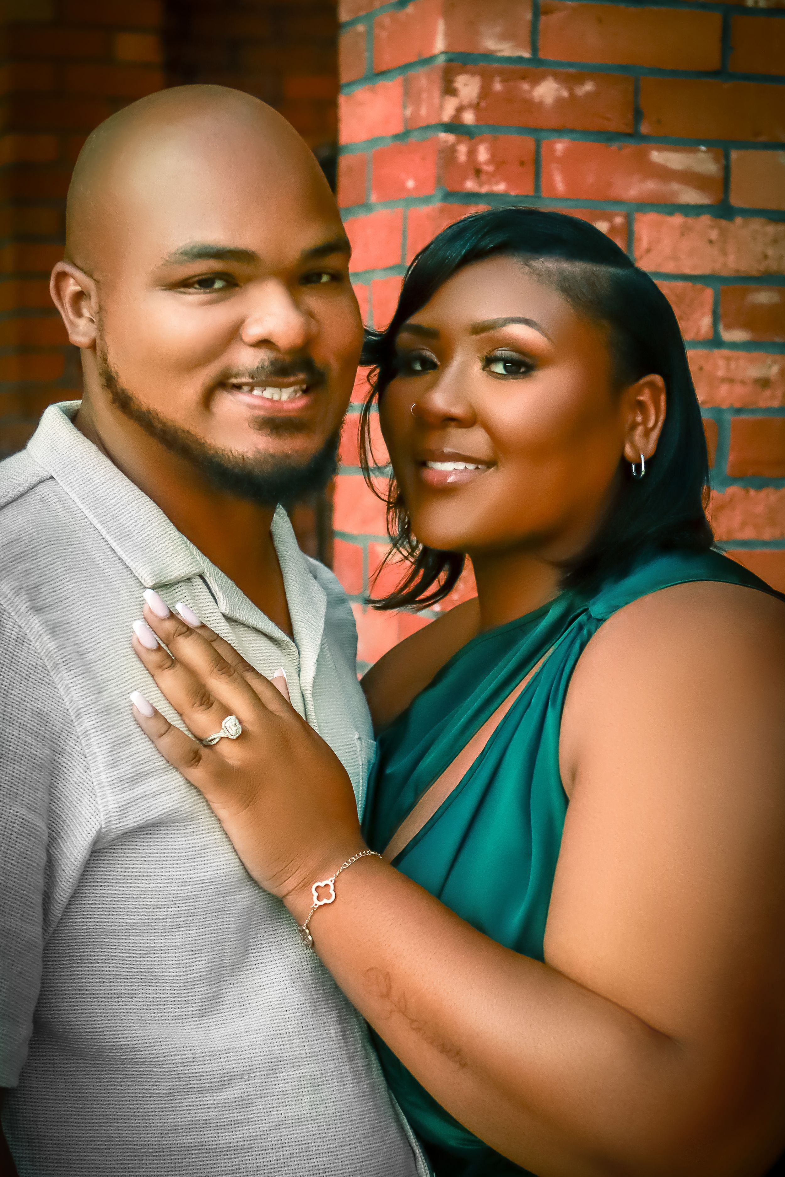 A happy couple poses against a brick wall backdrop, with one wearing a green dress and the other in a grey shirt.