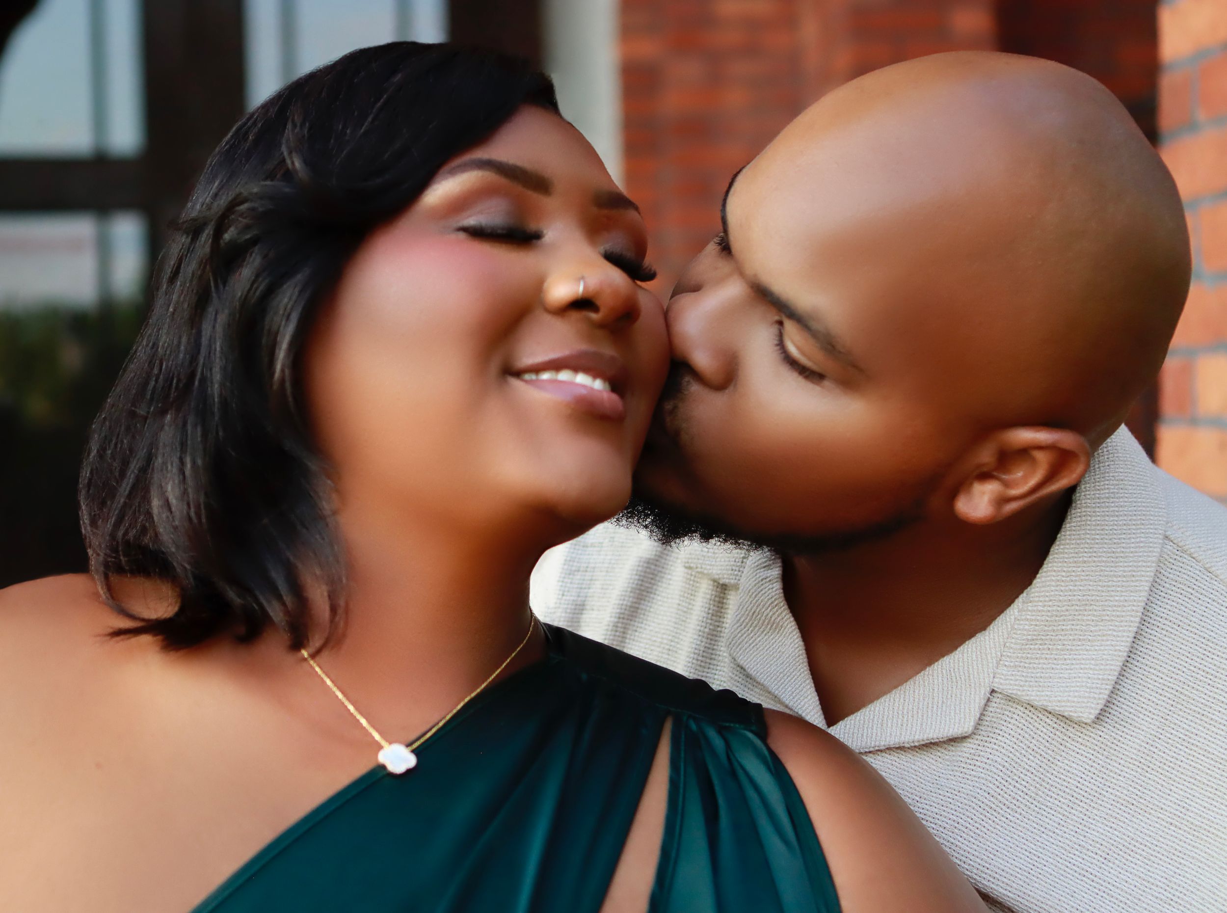 An affectionate couple sharing a tender moment together wearing casual attire against a brick background.