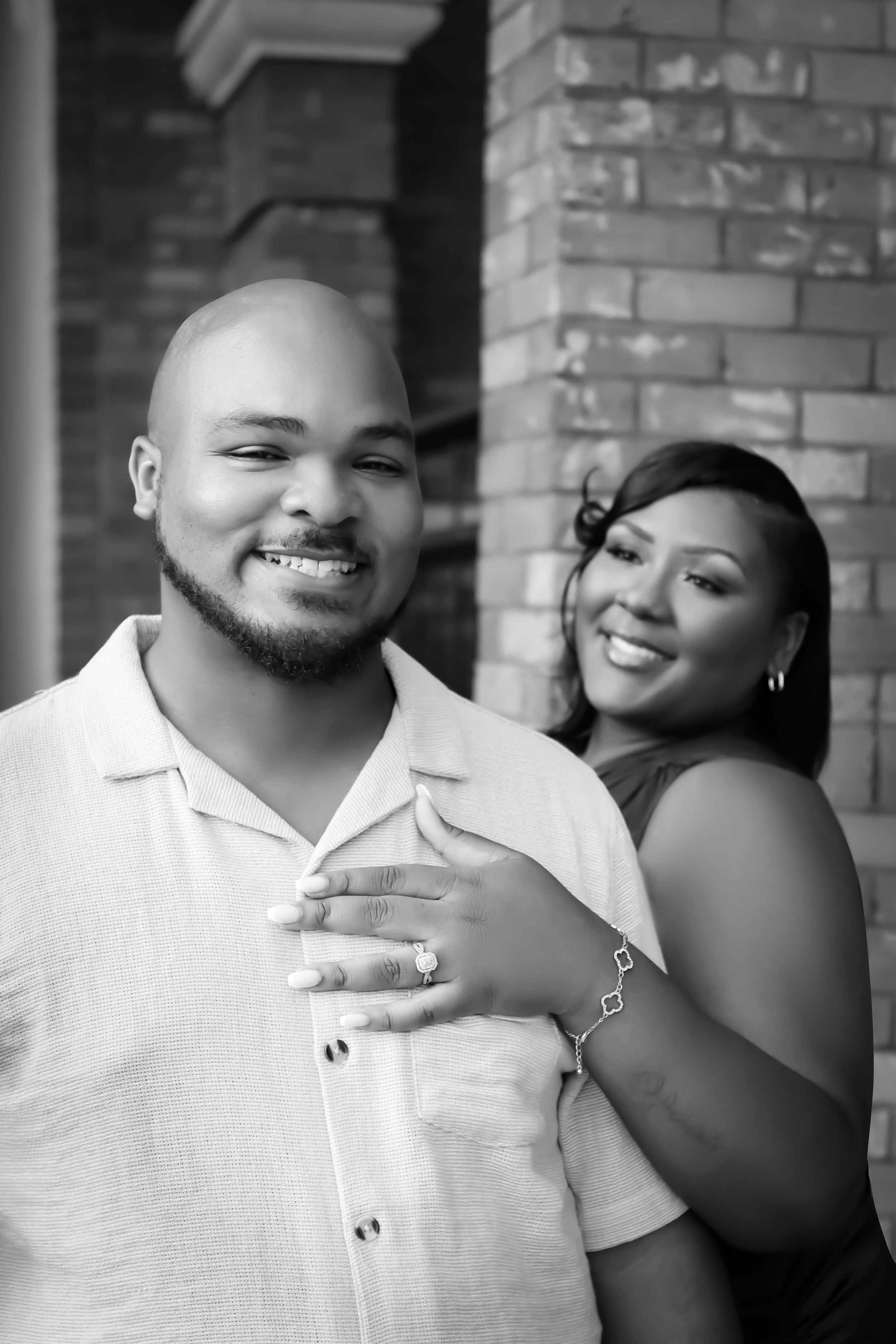 A happy couple embraces in a candid black and white portrait against a brick wall background.