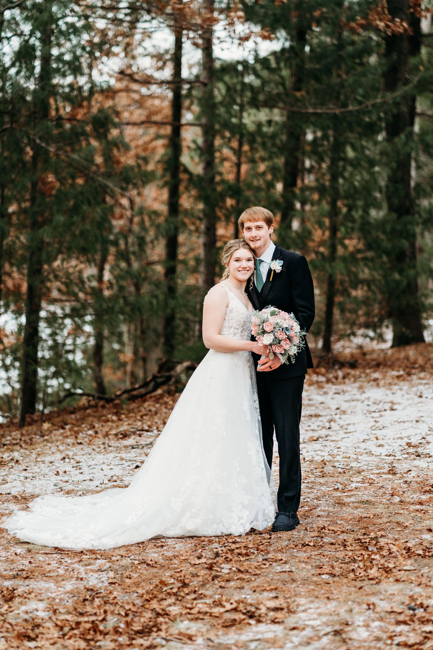 A couple in wedding attire poses on a leaf-covered path in an autumn forest.