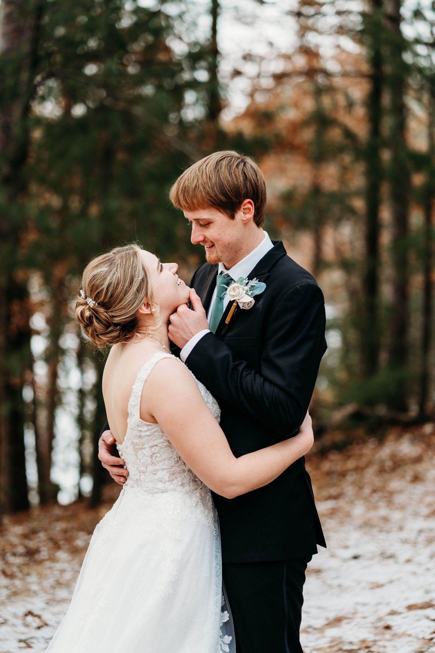 A newlywed couple embraces in a snowy winter forest setting during their wedding photoshoot.