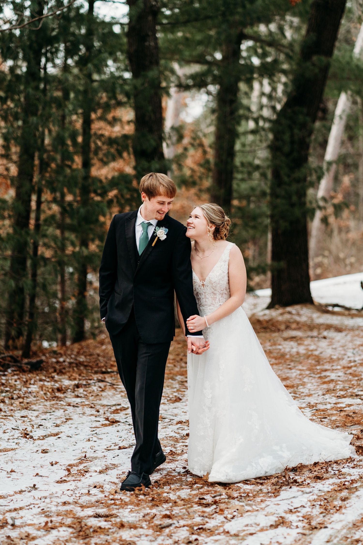 A bride and groom share a joyful moment walking together on a snowy forest path surrounded by tall trees in winter.