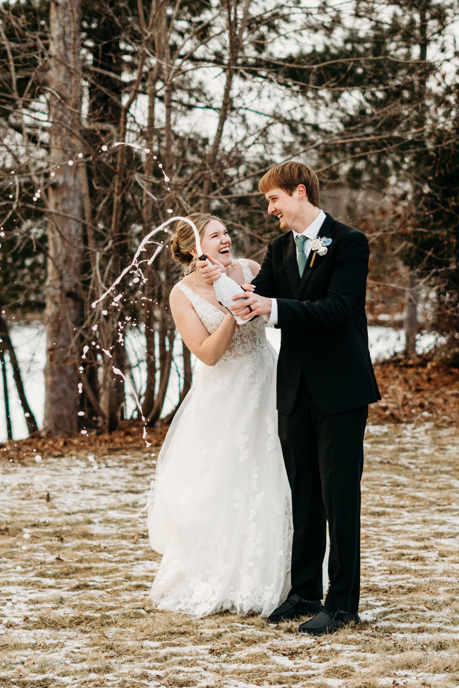 A wedding couple shares a joyful moment together in a snowy winter setting with pine trees in the background.