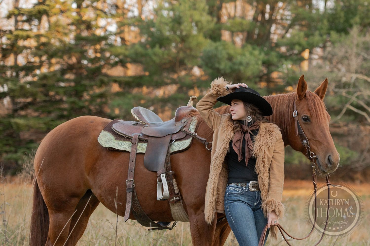 A brown horse with western saddle stands next to a figure in denim and tan leather jacket at sunset.