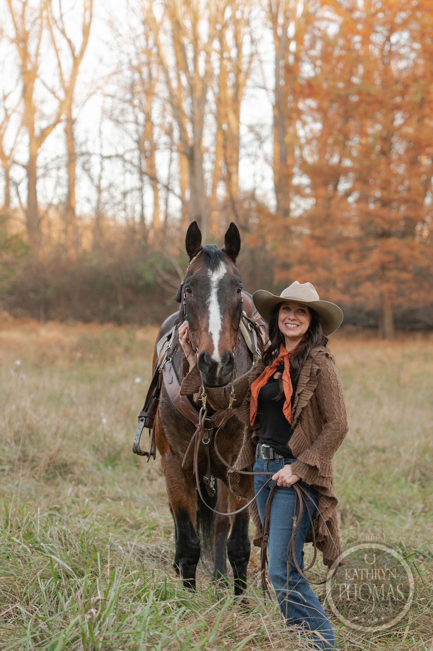 A horse and rider in western attire stand together in a golden field during autumn.